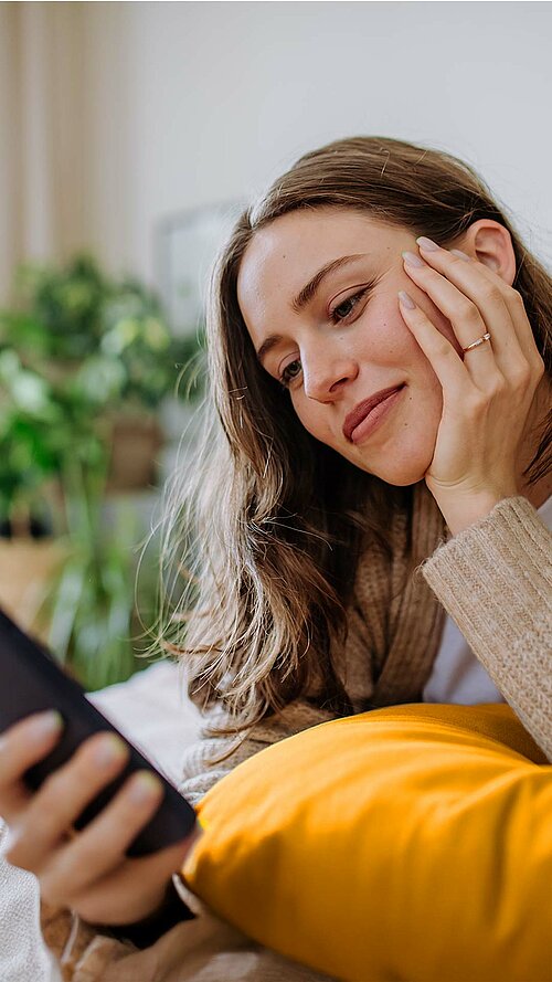 Person liegt auf einem Bett und schaut auf ein Smartphone, im Hintergrund sind Pflanzen und ein Fenster zu sehen