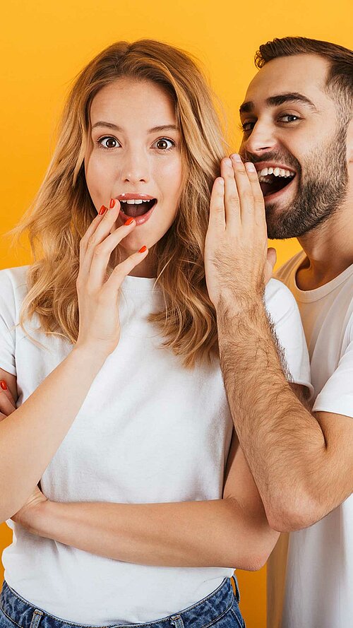 Zwei Personen stehen vor gelbem Hintergrund, eine Person flüstert der anderen etwas zu.
