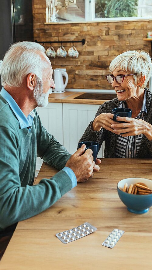 Älteres Paar sitzt am Küchentisch und trinkt Kaffee, mit Medikamentenblistern und einer Schüssel Kekse auf dem Tisch