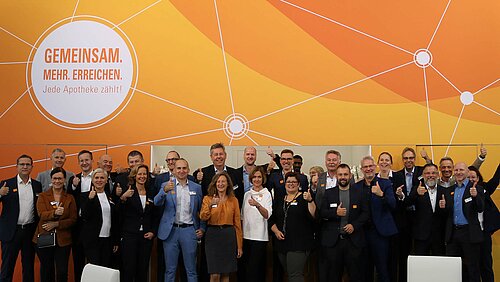 Gruppenfoto von ADG Mitarbeitern vor orangefarbener Wand mit dem Slogan ‚GEMEINSAM. MEHR. ERREICHEN. Jede Apotheke zählt!‘ – Symbolbild für Teamarbeit, Apothekennetzwerke und gemeinschaftliches Engagement im Gesundheitswesen