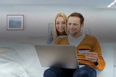 Paar auf weißem Sofa beim Online-Shopping, Mann hält Kreditkarte und nutzt Laptop, Frau schaut über seine Schulter