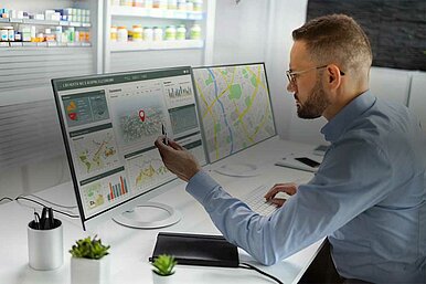 Mann analysiert Standortdaten auf zwei Monitoren in einem modernen Büro, mit Karten und Diagrammen zur Standortanalyse