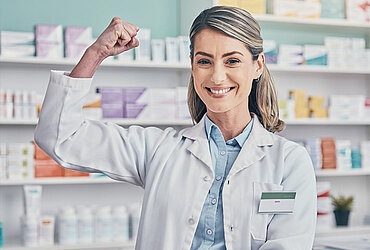 Lächelnde Apothekerin zeigt Stärke durch Pizeps-Geste in Apotheke