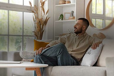 Mann sitzt entspannt auf einem modernen Sofa in einem hellen Wohnzimmer mit großen Fenstern und stilvoller Dekoration