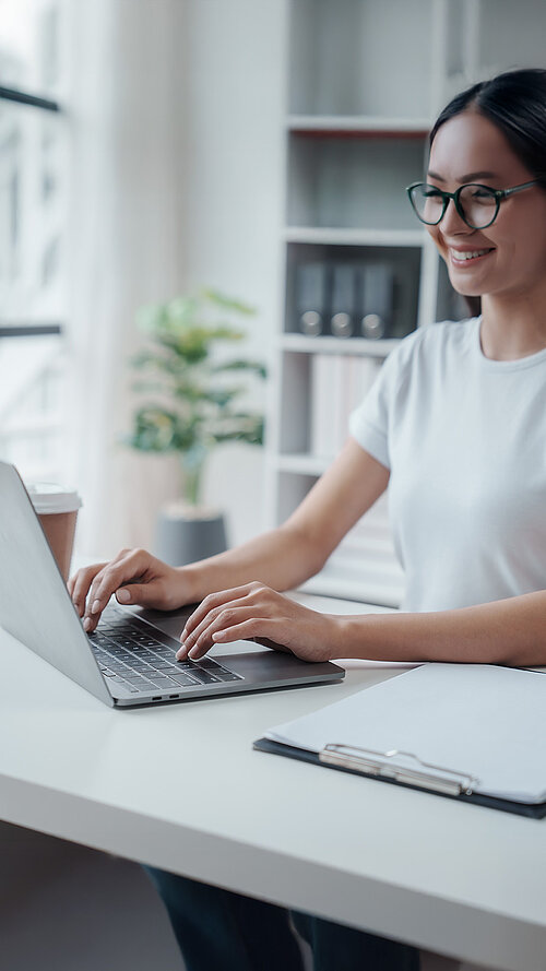 Person im Homeoffice an einem modernen Arbeitsplatz mit Laptop, Glas Wasser und Pflanzen im Hintergrund
