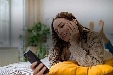 Person liegt auf einem Bett und schaut auf ein Smartphone, im Hintergrund sind Pflanzen und ein Fenster zu sehen