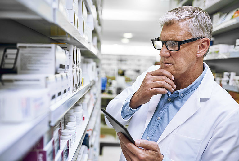 Apotheker mit Tablet-PC in der Hand schaut nachdenklich auf seine Ware im Regal